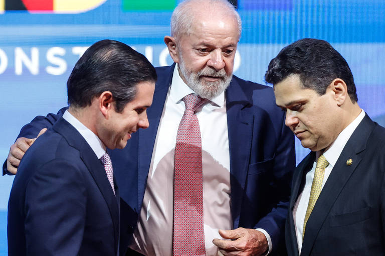 A imagem mostra três homens em um evento. O homem à esquerda está vestido com um terno escuro e uma gravata clara, enquanto o homem à direita usa um terno escuro com uma gravata amarela. O homem no centro, com cabelo grisalho e barba, está usando um terno escuro e uma gravata rosa. Eles parecem estar conversando e o homem do centro está segurando um papel.