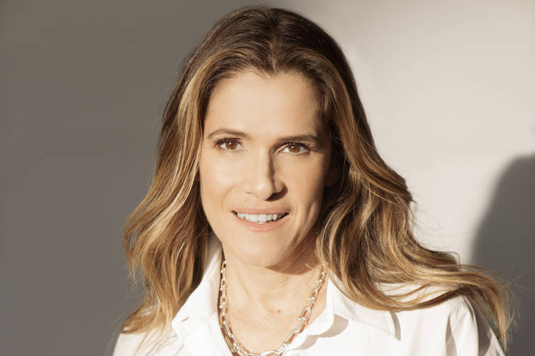 A imagem mostra uma mulher sorrindo, vestindo uma camisa branca de mangas longas, com a parte inferior da camisa levemente solta. Ela tem cabelos longos e ondulados, e está usando um colar prateado. A mulher está de pé, com as mãos nos bolsos de uma calça escura. O fundo é claro, com uma iluminação suave que destaca seu rosto.