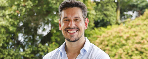 Renato (João Vicente de Castro) *** Local Caption *** Um homem está posando com os braços cruzados e sorrindo. Ele usa uma camisa de botão azul clara e calças marrons. Ao fundo, há uma vegetação densa e verde, com árvores e arbustos, criando um ambiente natural e agradável.