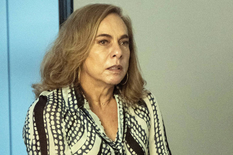 Uma mulher está em pé, apoiando-se em uma cadeira em um ambiente de escritório. Ela usa uma blusa com padrões em preto e branco e calças escuras. Seu cabelo é castanho claro e está solto. A expressão dela parece séria e concentrada. Ao fundo, há uma parede clara e uma porta.