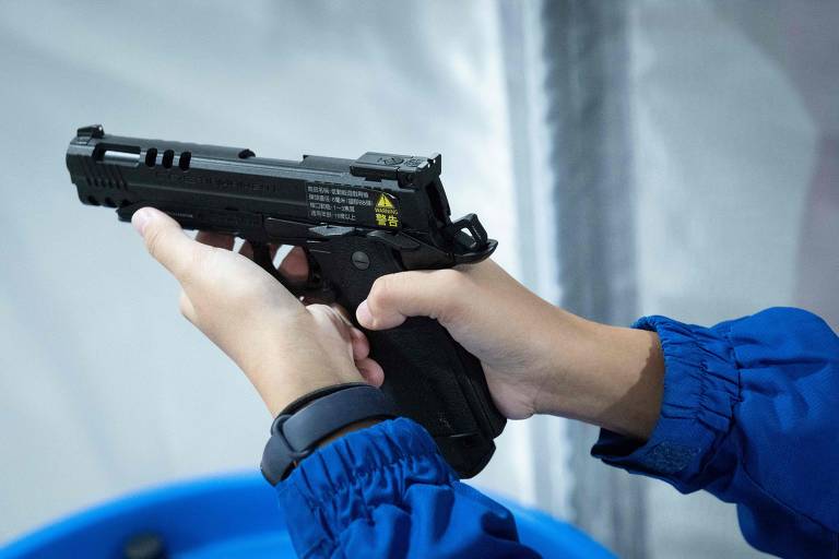 A imagem mostra uma pessoa segurando uma pistola com as duas mãos. A pistola é de cor preta e parece ser uma réplica ou uma arma de treinamento. A pessoa está usando uma camisa de manga longa azul e um relógio no pulso. Ao fundo, há um recipiente azul.