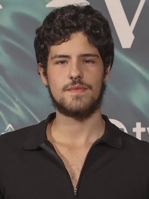 Um homem jovem está posando em um evento, vestindo uma camisa preta de gola e calças escuras. Ele tem cabelo escuro e barba. Ao fundo, há um painel com as palavras 'MANIA' e 'VOCÊ', além do logotipo da TV Globo.