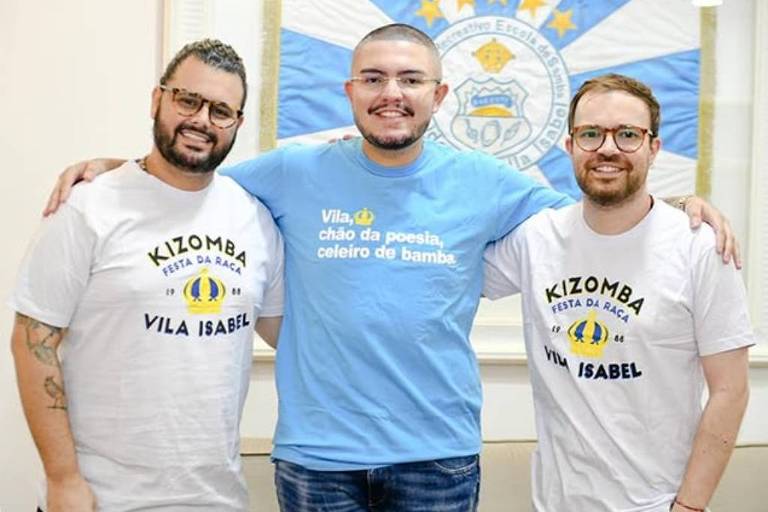 Três homens estão posando juntos em um ambiente interno. O homem à esquerda usa uma camiseta branca com a inscrição 'KIZOMBA FESTA DA RAÇA VILA ISABEL' e tem barba e óculos. O homem do meio está usando uma camiseta azul com a frase 'Vila, chão da poesia, celeiro de bamba' e tem cabelo curto e óculos. O homem à direita também usa uma camiseta branca com a mesma inscrição que o da esquerda e tem barba e óculos. Ao fundo, há uma bandeira com cores azul e branca.