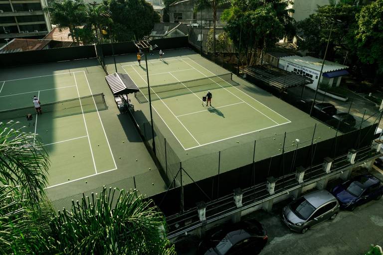 A imagem mostra um complexo de quadras de tênis. Há duas quadras visíveis, com jogadores em ação. Uma das quadras possui uma cobertura ao lado. Ao fundo, há prédios altos e árvores ao redor. Na parte inferior da imagem, é possível ver alguns carros estacionados.
