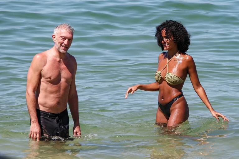 Um homem e uma mulher estão na água do mar. O homem é de pele clara, sem camisa, com cabelo grisalho e usa calças escuras. A mulher tem pele morena, cabelo cacheado e usa um biquíni verde. Ambos parecem estar se divertindo na água.