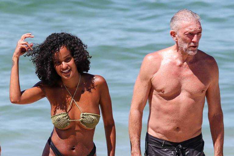 Um homem e uma mulher caminham na praia. A mulher tem cabelo cacheado e usa um biquíni verde, enquanto o homem está sem camisa e usa shorts de banho escuros. Eles estão saindo da água e a praia tem areia clara e ondas ao fundo.