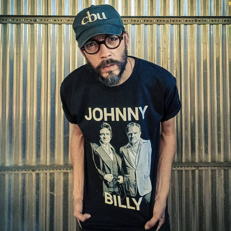 Um homem com barba e óculos está posando em frente a um fundo metálico. Ele usa uma camiseta preta com a inscrição 'JOHNNY BILLY' e uma imagem em preto e branco de duas pessoas. O homem também está usando um boné cinza com a inscrição 'cbu'.