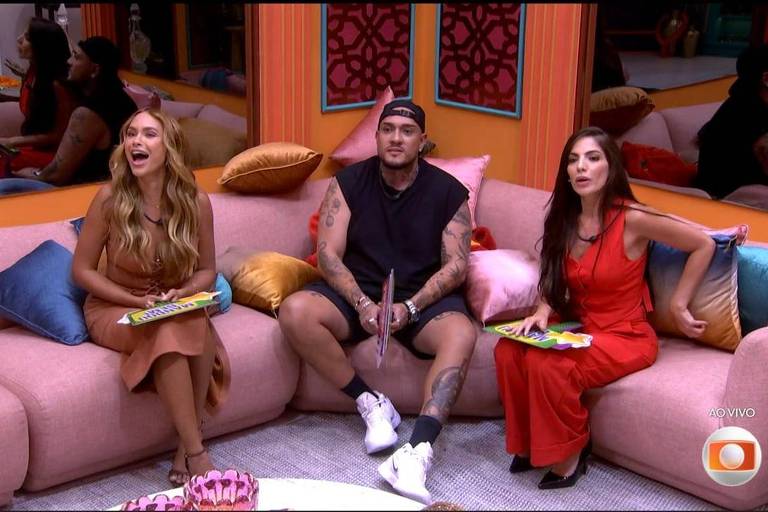 A imagem mostra três pessoas sentadas em um sofá rosa em um ambiente interno. À esquerda, uma mulher com cabelo longo e liso, vestindo um vestido marrom, está sorrindo. No centro, um homem com cabelo curto e tatuagens, usando uma camiseta preta e shorts, está segurando um objeto. À direita, uma mulher com cabelo longo e liso, vestindo um vestido vermelho, também está sorrindo. Ao fundo, outras pessoas podem ser vistas, mas não estão claramente focadas.