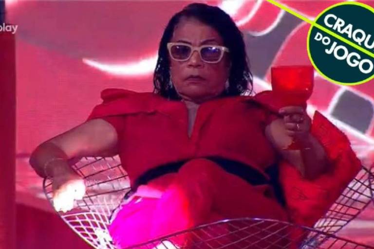 A imagem mostra uma mulher sentada em uma cadeira de design moderno, com um vestido vermelho e óculos escuros. Ela segura uma taça vermelha e tem uma expressão séria. O fundo é decorado em tons de vermelho e rosa, com luzes brilhantes. No canto superior direito, há um selo que diz 'CRAQUE DO JOGO'.