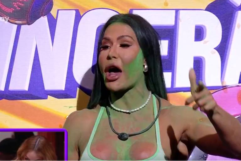 Uma mulher com cabelo longo e escuro, usando um top verde claro, está falando em um palco. Seu rosto está parcialmente coberto com tinta verde. Ao fundo, há um grande letreiro com a palavra 'SINGEIRA' em letras grandes e coloridas. Na parte inferior da imagem, há uma pequena janela com outra pessoa visível.