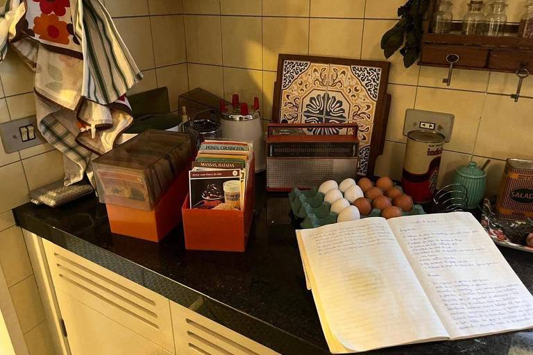 A imagem mostra uma bancada de cozinha com diversos itens. Há caixas organizadoras em laranja, um caderno aberto com anotações, uma bandeja com ovos brancos e marrons, e um rádio antigo. Ao fundo, há uma toalha pendurada e uma decoração de azulejos. A parede é revestida com azulejos claros.