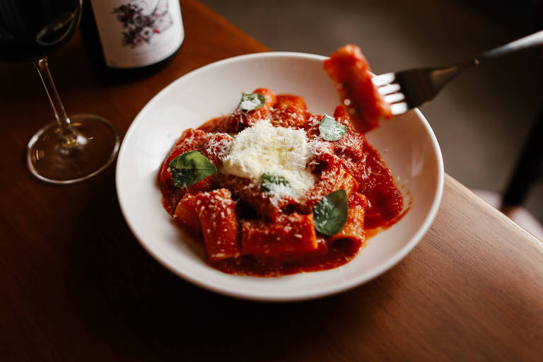 Um prato redondo branco contendo massa tipo rigatoni coberta com molho de tomate, folhas de manjericão e queijo parmesão ralado. Um garfo está prestes a pegar um pedaço da massa. Ao lado, há uma taça de vinho tinto e uma garrafa de vinho parcialmente visível ao fundo.