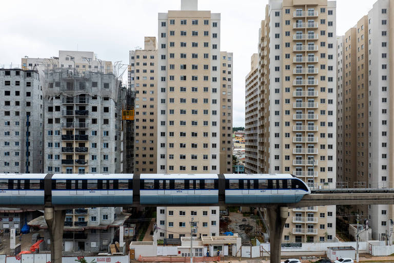 A imagem mostra um trem elevado passando por uma área urbana em construção, cercado por prédios altos. À esquerda, há um edifício em construção com andaimes, enquanto à direita, há vários prédios residenciais com janelas. O céu está nublado, e a cena transmite uma sensação de desenvolvimento urbano.