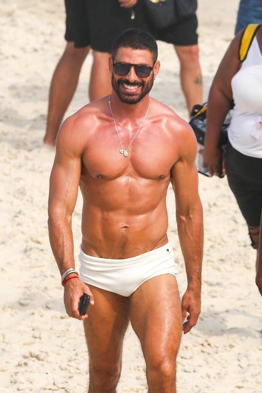 Um homem musculoso caminha na praia, usando um sunga branca e óculos escuros. Ele tem um sorriso no rosto e está bronzeado. Ao fundo, há outras pessoas na praia, algumas com roupas de banho e outras com roupas casuais.