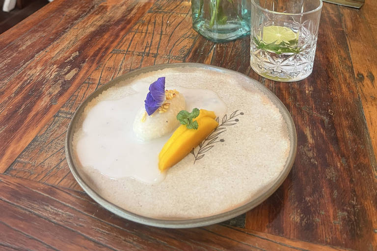 A imagem mostra uma mesa de restaurante com um prato decorativo contendo uma porção de comida, que inclui uma fatia de manga e um pequeno montinho de arroz, decorado com uma flor roxa e um trevo. Ao lado, há um copo com uma bebida e uma garrafinha de vidro com flores coloridas, incluindo flores laranjas e brancas. O ambiente é acolhedor, com paredes amarelas e mesas de madeira.