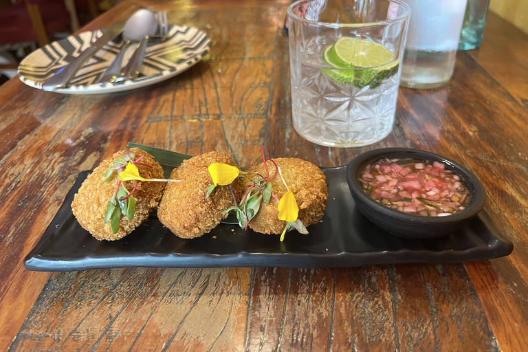 A imagem mostra um prato preto com três croquetes empanados, decorados com pequenas flores amarelas e folhas verdes. Ao lado, há um pequeno recipiente com molho, que contém cebolas picadas. Ao fundo, uma bebida em um copo transparente com uma fatia de limão e gelo, além de um prato com talheres em uma mesa de madeira.