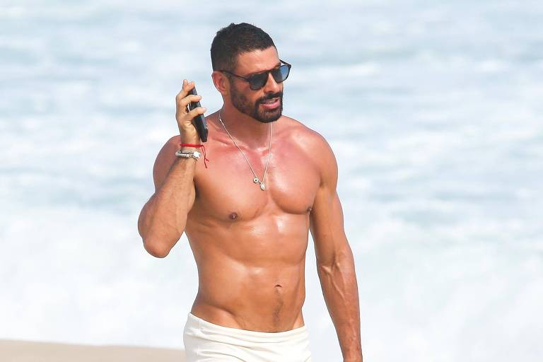 Um homem musculoso caminha na praia, usando apenas um sunga branca. Ele está descalço e segurando um celular, com óculos de sol e um colar. Ao fundo, vê-se o mar e a areia da praia.