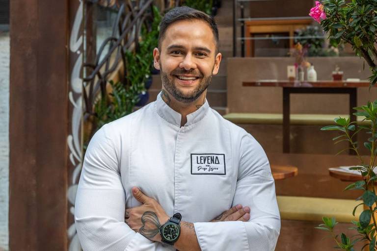 Um homem sorridente está posando em frente a uma confeitaria chamada 'Café da Manhã'. Ele usa um uniforme de chef branco e tem um relógio no pulso. Ao fundo, há uma decoração com plantas e mesas, além de uma placa com o nome da confeitaria.
