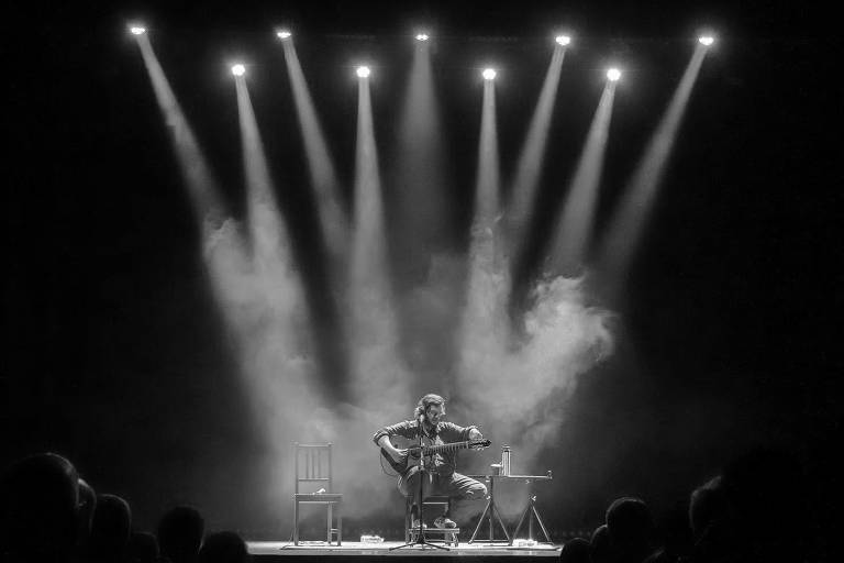 A imagem mostra um artista sentado em um palco, tocando violão. Ele está iluminado por vários holofotes que criam um efeito dramático com fumaça ao fundo. Duas cadeiras estão visíveis ao lado do artista, e a plateia é parcialmente visível na parte inferior da imagem, em um ambiente escuro.