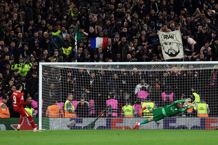 A imagem captura um momento de um jogo de futebol, onde um jogador, vestido com um uniforme vermelho, está prestes a chutar um pênalti. O goleiro, vestido de verde, está se lançando para o lado direito em uma tentativa de defender a cobrança. Ao fundo, uma multidão de torcedores está visível, com alguns segurando bandeiras e cartazes, incluindo um que diz 'URBAN'. A atmosfera é intensa, com segurança visível na área.