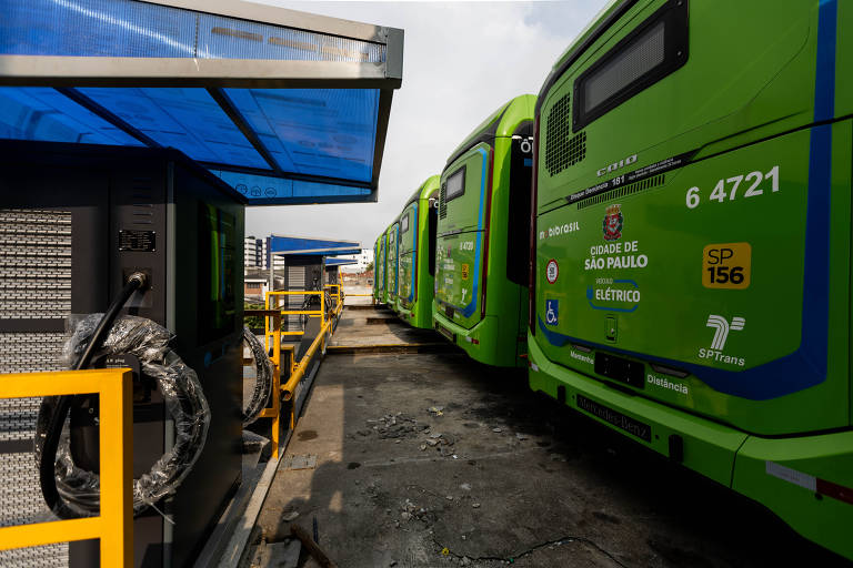 A imagem mostra uma estação de recarga para ônibus elétricos na cidade de São Paulo. Vários ônibus verdes estão alinhados, com um deles visível em primeiro plano, mostrando o número 6 4721. Ao lado, há um equipamento de recarga com cabos conectados. O céu está claro e há uma cobertura azul sobre a estação