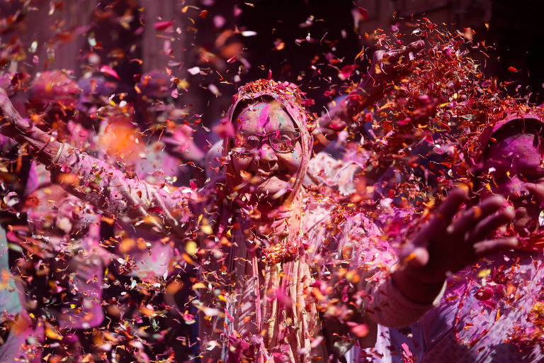 A imagem mostra uma celebração do festival Holi, com pessoas sorrindo e dançando enquanto são cobertas por pétalas coloridas. Uma figura central, com rosto pintado e vestimentas tradicionais, levanta os braços em celebração, cercada por uma chuva de flores. O ambiente é vibrante e festivo, com uma mistura de cores e alegria.