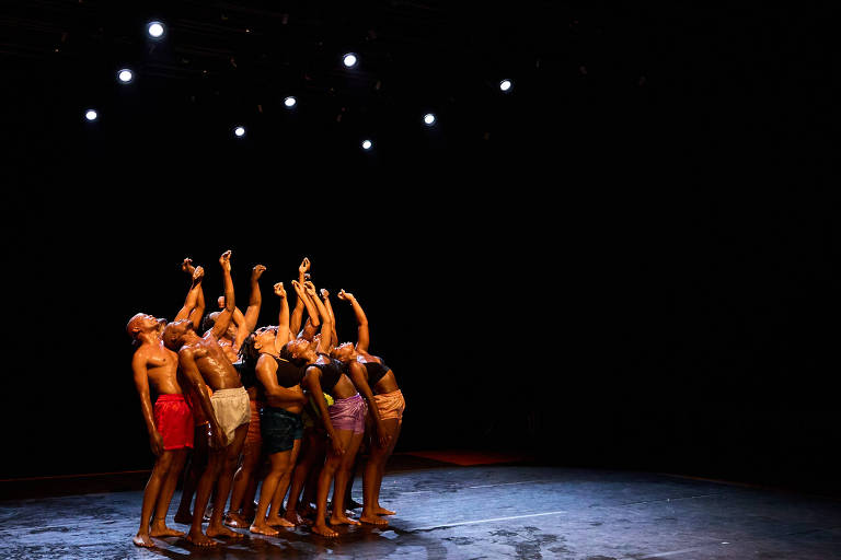 Um grupo de dançarinos se apresenta em um palco escuro, com luzes focadas sobre eles. Os dançarinos estão em uma formação em que se inclinam para trás, com os braços estendidos para cima. Eles usam roupas de dança coloridas e estão descalços. O chão do palco reflete a luz, criando um efeito visual interessante.