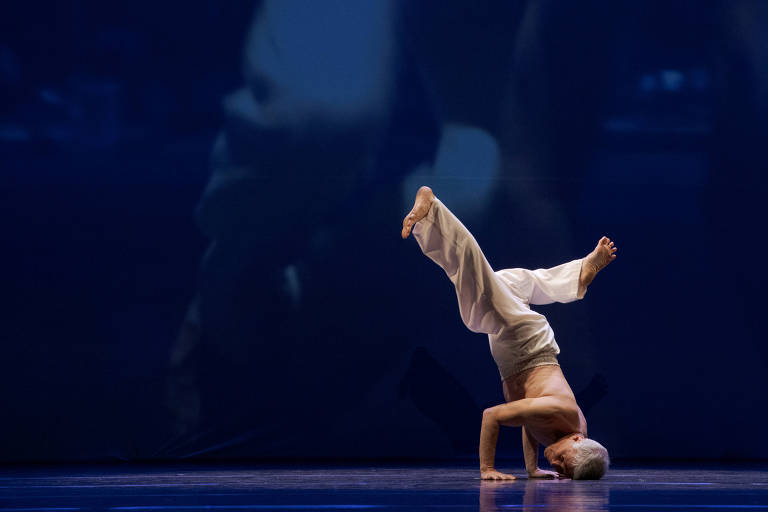 Um dançarino está realizando uma manobra acrobática em um palco. Ele está apoiado em uma mão, com uma perna estendida para cima e a outra perna dobrada. O dançarino é idoso e usa uma roupa leve e clara. O fundo é de um tom azul escuro, com uma projeção sutil que sugere movimento.