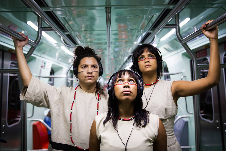 Três pessoas estão dentro de um metrô, segurando nas barras de apoio. Elas usam fones de ouvido e vestem roupas simples, com colares de contas. A mulher no centro tem cabelo liso e usa um colar com contas vermelhas e brancas, enquanto os outros dois têm cabelo encaracolado e liso, respectivamente. O ambiente é moderno, com paredes e assentos em tons de verde claro.