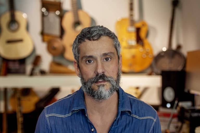 homem branco de barba e cabelo grisalhos encara a câmera diante de vários violões e guitarras