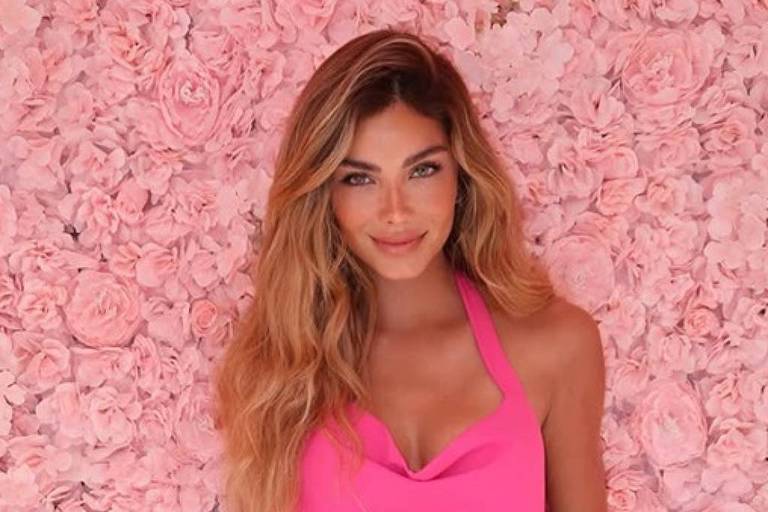 A imagem mostra uma mulher posando em frente a um fundo de flores rosas. Ela está usando um vestido rosa justo, com alças finas e um decote em forma de coração. Seu cabelo é longo e ondulado, e ela sorri para a câmera.