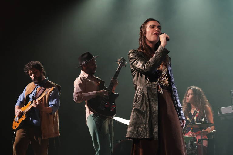 A imagem mostra uma apresentação musical ao vivo com quatro músicos no palco. Uma mulher está cantando ao microfone, vestindo um casaco de couro e uma saia longa. À sua esquerda, um homem toca guitarra elétrica, enquanto um segundo homem, ao fundo, toca um baixo. À direita, uma mulher está sentada atrás de uma bateria, observando a performance. O ambiente é iluminado com luzes suaves, criando uma atmosfera de show.