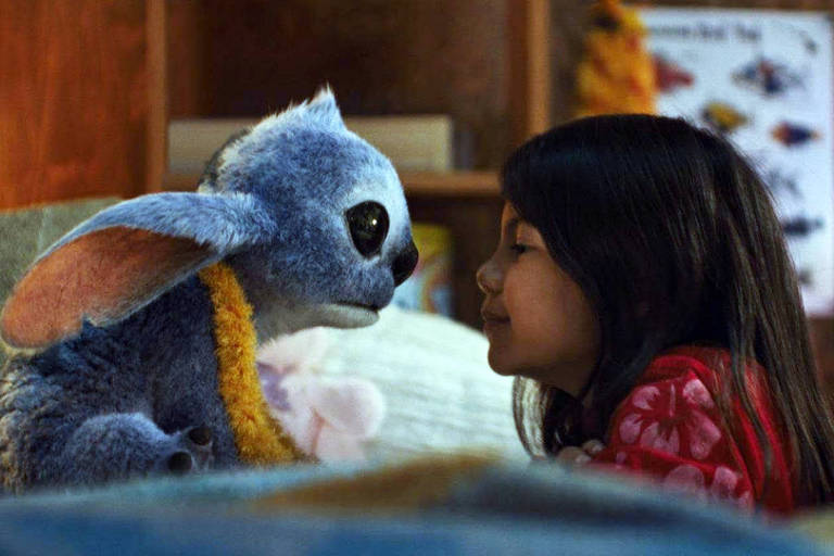 Cena do filme Lilo & Stitch - Live Action (2025) *** Local Caption *** A imagem mostra uma criança com cabelo longo e escuro, usando uma blusa vermelha, olhando para um boneco de pelúcia que se assemelha a um alienígena azul com grandes orelhas. Eles estão próximos um do outro, em um ambiente interno que parece ser um quarto, com prateleiras ao fundo e alguns objetos decorativos visíveis.