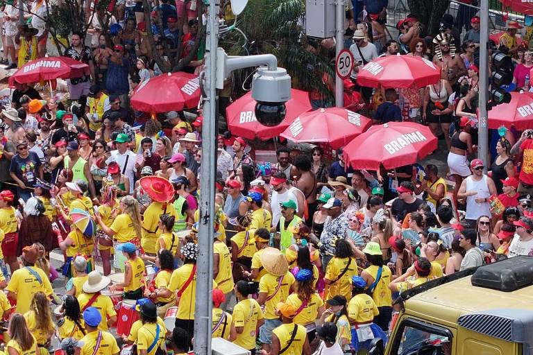 A imagem mostra uma grande multidão reunida durante uma festividade, possivelmente o Carnaval. As pessoas estão vestidas com camisetas amarelas e muitos usam chapéus coloridos. Há várias sombrinhas vermelhas com a marca 'Brahma' ao fundo. A cena é animada, com pessoas dançando e se divertindo. Um veículo amarelo está visível na parte inferior da imagem.