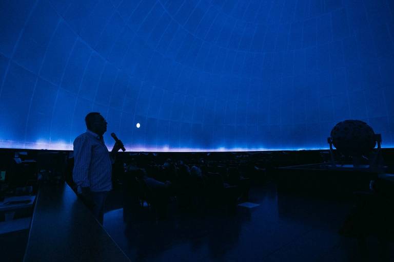 A imagem mostra um interior de um planetário com iluminação azul. Um homem está de perfil, segurando um microfone, enquanto fala para uma audiência visível em cadeiras. No fundo, há uma representação da lua projetada na cúpula do planetário, e um modelo de corpo celeste está posicionado à direita.