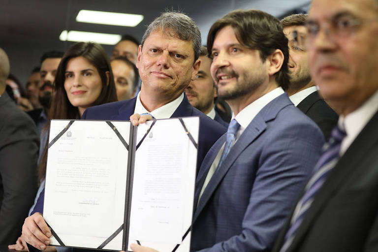 A imagem mostra um grupo de pessoas em um ambiente interno, possivelmente em um evento oficial. Dois homens estão em destaque, um deles segurando um documento assinado. O homem à esquerda tem cabelo curto e usa um terno escuro, enquanto o homem à direita tem cabelo mais longo e usa um terno claro. Ao fundo, há outras pessoas observando a cena, incluindo uma mulher com cabelo longo e escuro. O ambiente parece ser bem iluminado.