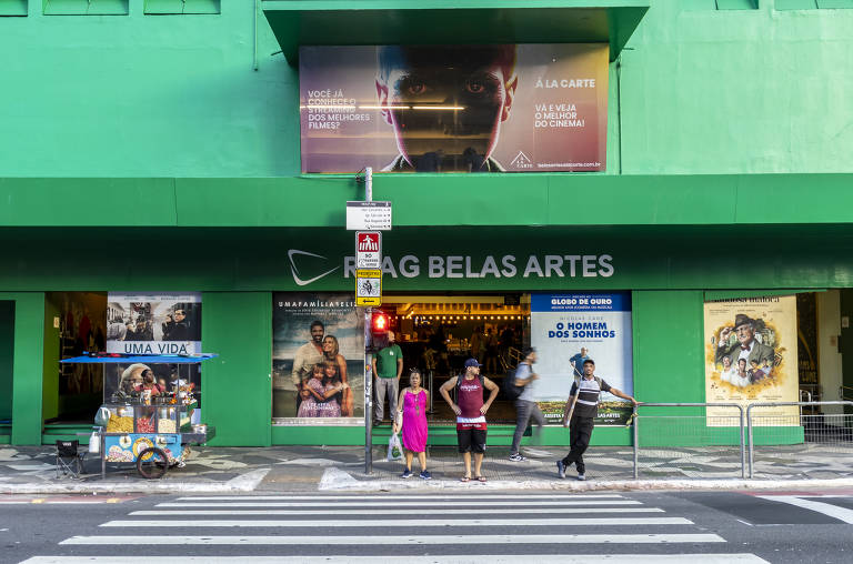 A imagem mostra a fachada do cinema Belas Artes, que é pintada de verde. Na parte superior, há um grande cartaz com a imagem de um homem. Na entrada, há pessoas esperando para atravessar a rua, e um carrinho de vendas está estacionado na calçada. O sinal de trânsito está vermelho. Várias cartazes de filmes estão visíveis nas paredes do cinema.