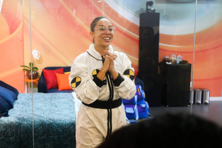Uma mulher está em um ambiente de vidro, vestindo um macacão branco com detalhes em preto e amarelo. Ela está sorrindo e com as mãos juntas, em uma expressão de alegria ou expectativa. Ao fundo, há uma cama com lençóis azuis e almofadas, além de uma decoração colorida nas paredes. O ambiente parece ser parte de um reality show.