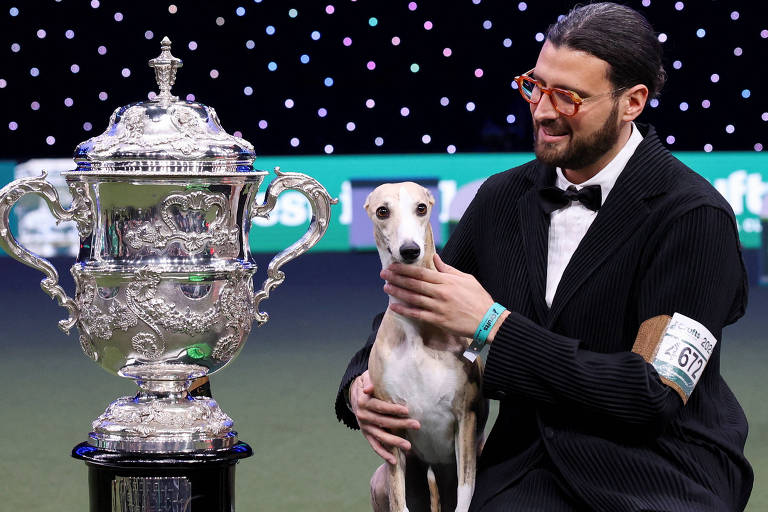 Um homem com óculos e um terno preto está sentado ao lado de um cão da raça Whippet. Ele sorri enquanto acaricia o cão, que está em pé ao seu lado. À esquerda, há um grande troféu prateado em cima de uma base preta, e à direita, um prêmio verde. O fundo é decorado com luzes brilhantes.