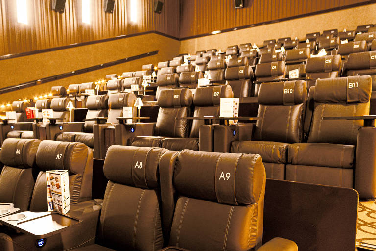 A imagem mostra uma sala de cinema com poltronas reclináveis em couro marrom. As poltronas estão dispostas em fileiras, com mesas laterais para bebidas ou alimentos. O ambiente é bem iluminado, com luzes suaves nas paredes e um padrão de carpete no chão. Não há pessoas visíveis na imagem.