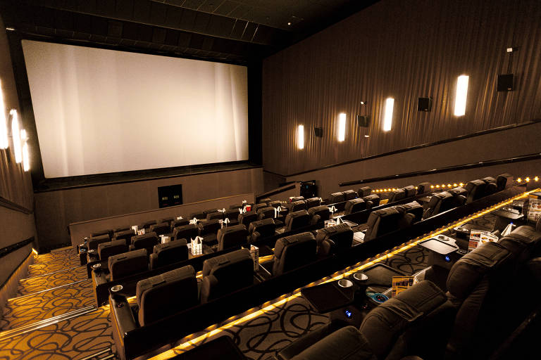 A imagem mostra o interior de uma sala de cinema. Há um grande telão em branco na frente, com assentos confortáveis dispostos em fileiras. As paredes são de um tom escuro e há iluminação suave nas laterais. O chão possui um padrão decorativo e há alguns itens de cinema, como copos e embalagens de pipoca, espalhados pelos assentos.