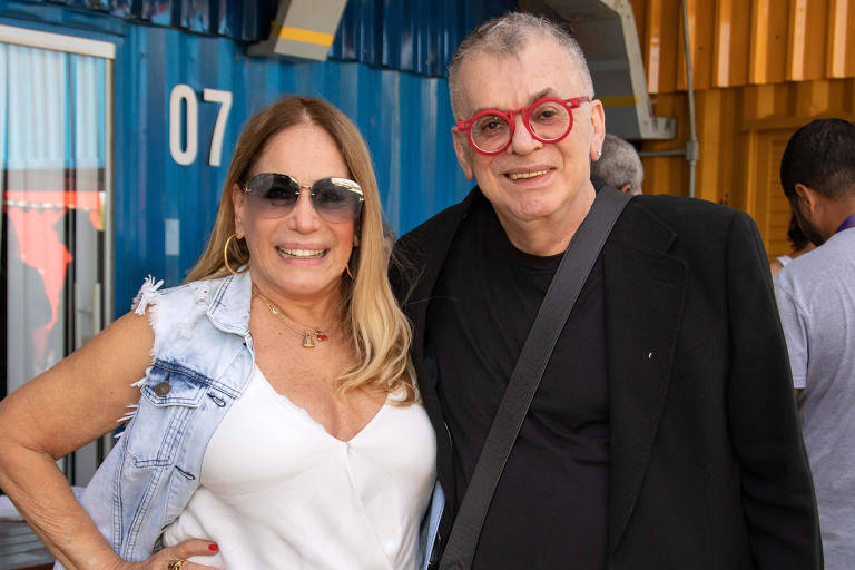 A imagem mostra duas pessoas posando para a foto em um ambiente ao ar livre. A mulher à esquerda usa uma blusa branca e um colete jeans, com óculos escuros e cabelo solto. O homem à direita veste uma camisa preta e um blazer, com óculos de armação vermelha. Ao fundo, há containers coloridos e pessoas em pé, algumas delas visíveis.