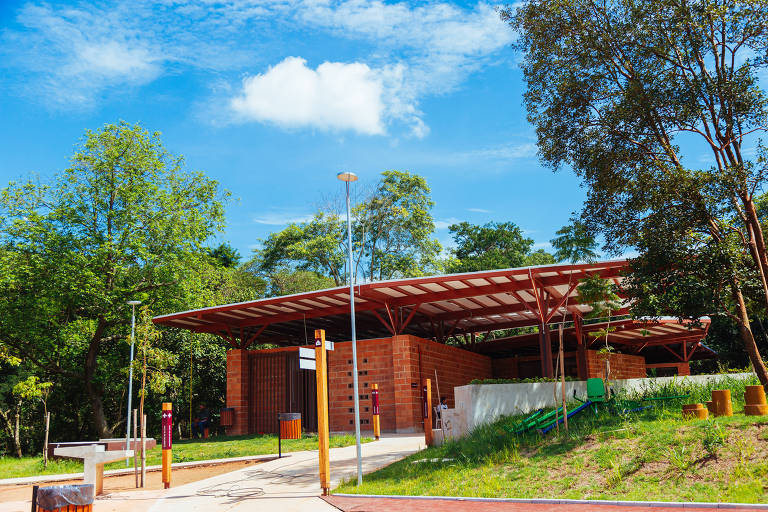 A imagem mostra um edifício moderno com estrutura de telhado em forma de cobertura, localizado em uma área verde. O edifício é feito de tijolos e possui janelas amplas. Ao redor, há árvores e um gramado bem cuidado. O céu está claro com algumas nuvens brancas.