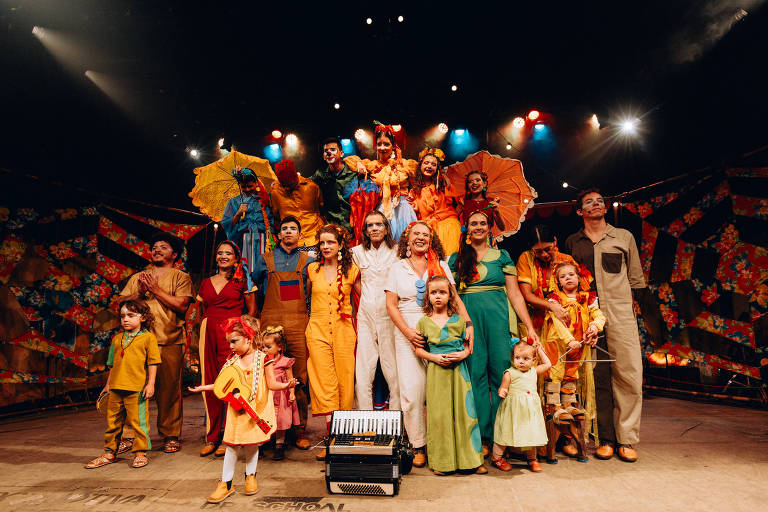 Um grupo de pessoas em um palco, posando para a foto. O elenco é composto por adultos e crianças, todos vestidos com trajes coloridos e variados. Algumas pessoas seguram guarda-chuvas coloridos, enquanto outras estão em poses alegres. Ao centro, há um acordeão no chão. O fundo é decorado com padrões vibrantes.