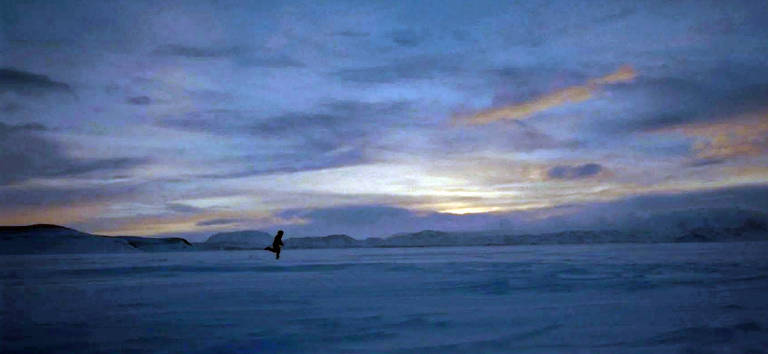 A imagem mostra uma vasta paisagem coberta de neve sob um céu azul escuro com nuvens. No centro, uma figura humana caminha sozinha sobre a neve, com montanhas ao fundo. O céu apresenta tons de azul e laranja, indicando o entardecer