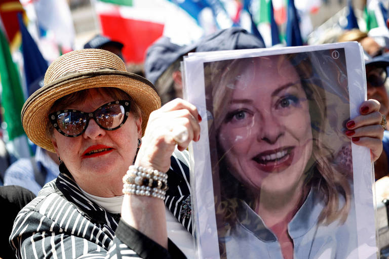 Uma mulher com um chapéu de palha e óculos escuros segura um cartaz com a imagem de uma mulher sorridente. Ela está em meio a uma multidão, com bandeiras ao fundo. A mulher usa uma blusa listrada e pulseiras. O ambiente parece ser de uma manifestação ou evento público.