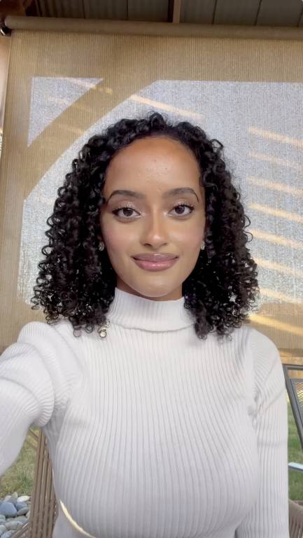 A imagem mostra uma mulher com cabelo cacheado e escuro, usando um suéter branco de gola alta. Ela está sorrindo levemente e olhando para a câmera. Ao fundo, há uma cortina translúcida que permite a entrada de luz natural, criando um ambiente iluminado.