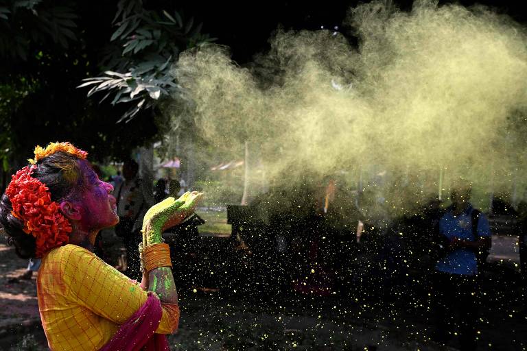 Uma mulher com a pele colorida em tons de rosa e verde, usando um vestido amarelo e adornos florais na cabeça, está soprando pó colorido no ar durante a celebração do Holi. O ambiente é ao ar livre, com árvores ao fundo e pessoas visíveis em segundo plano.