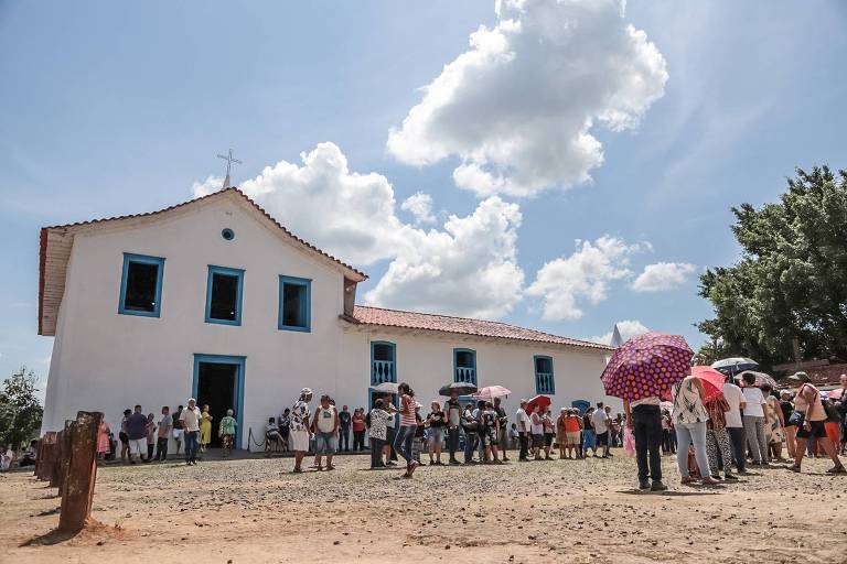 A imagem mostra uma fila de pessoas em frente a uma igreja branca com janelas azuis e um telhado de telhas vermelhas. As pessoas estão usando guarda-sóis coloridos, e o céu está parcialmente nublado com algumas nuvens. O ambiente é rural, com um chão de terra e algumas árvores ao fundo.