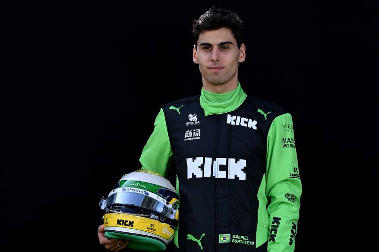 Um jovem piloto de Fórmula 1 está posando para a foto. Ele usa um macacão preto com detalhes em verde e a palavra 'KICK' em destaque. O piloto segura um capacete de corrida que possui cores verde, amarelo e azul, refletindo as cores da bandeira do Brasil. O fundo da imagem é escuro, destacando o piloto e seu equipamento.